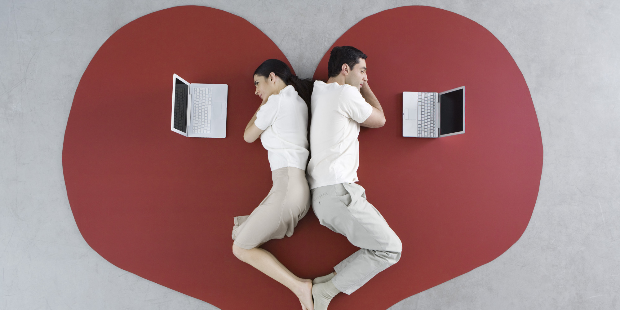 Man and woman lying back to back on large heart, both looking at laptop computers