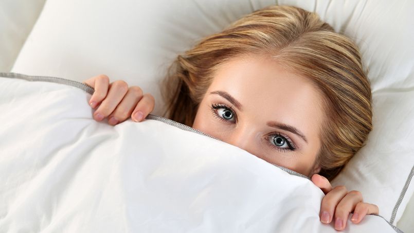 Beautiful blonde woman hiding face under cover lying in bed. Female sparky eyes looking in camera closeup. Sweet dreams, flirtation, playing game, wake up in strange place, shame, casual sex concept